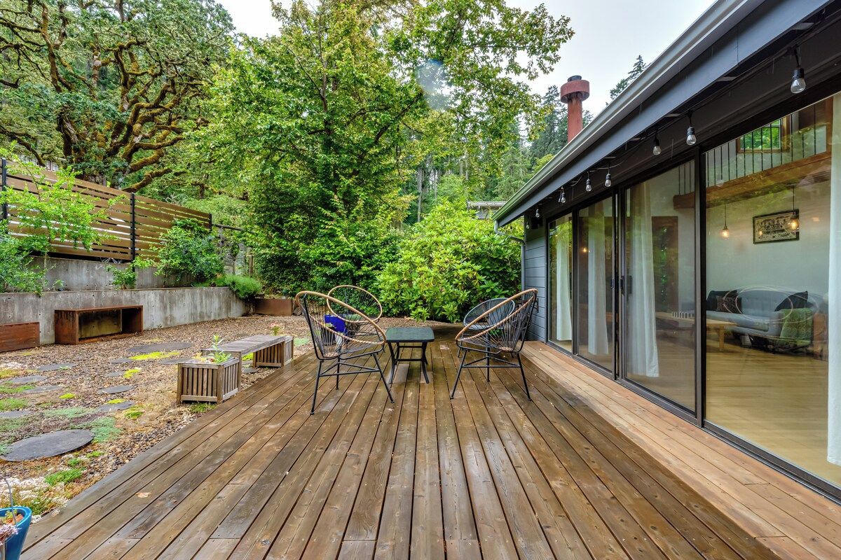 Eine Holzterrasse mit Tisch und Stühlen, umgeben von Grünpflanzen und einem Gebäude mit großen Fenstern.