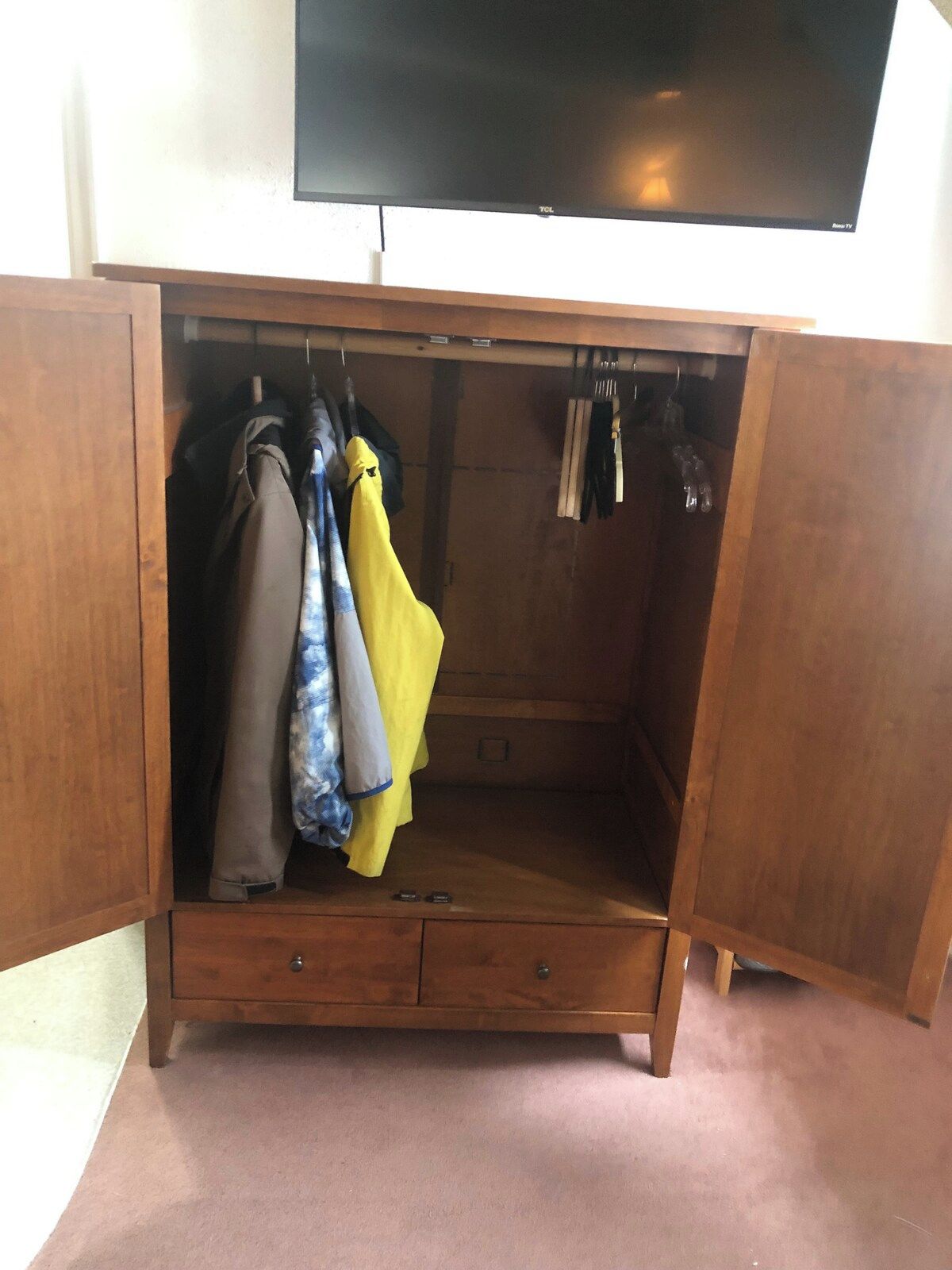 A wooden wardrobe with a television above it, containing hanging clothes and a small drawer.
