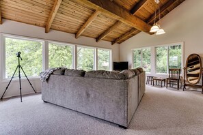 A room with a large gray sofa, a wooden ceiling, and large windows with a view of trees. - Waterfront House w/ Private Dock & Game Room (Veneta)