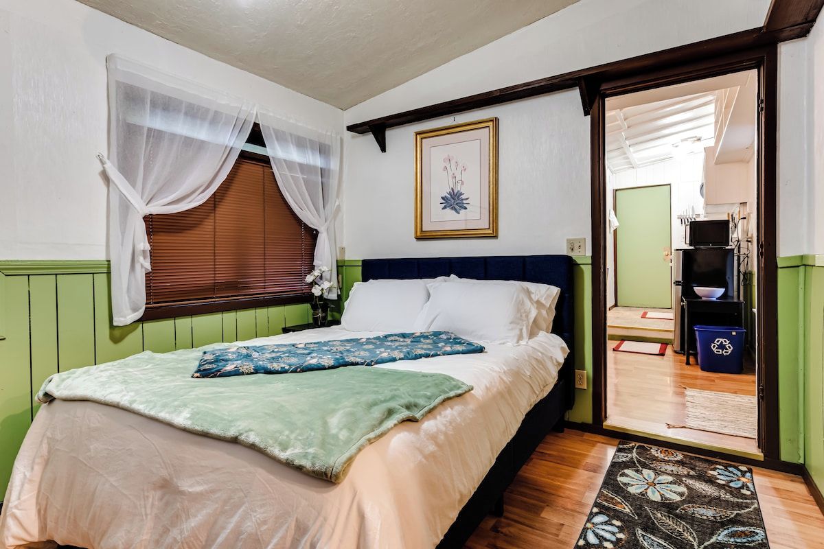 Une chambre à coucher avec un lit, une fenêtre ornée de rideaux légers, un tableau encadré au mur et une porte donnant sur une salle de bain.