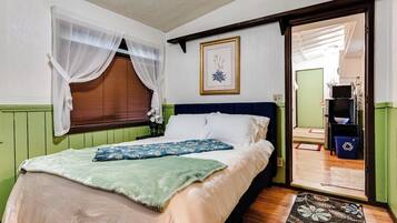 A bedroom with a bed, a window with sheer curtains, a framed picture on the wall, and a door leading to a bathroom.
