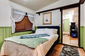 Ein Schlafzimmer mit einem Bett, einem Fenster mit leichtem Vorhang, einem gerahmten Bild an der Wand und einer Tür zu einem Badezimmer.