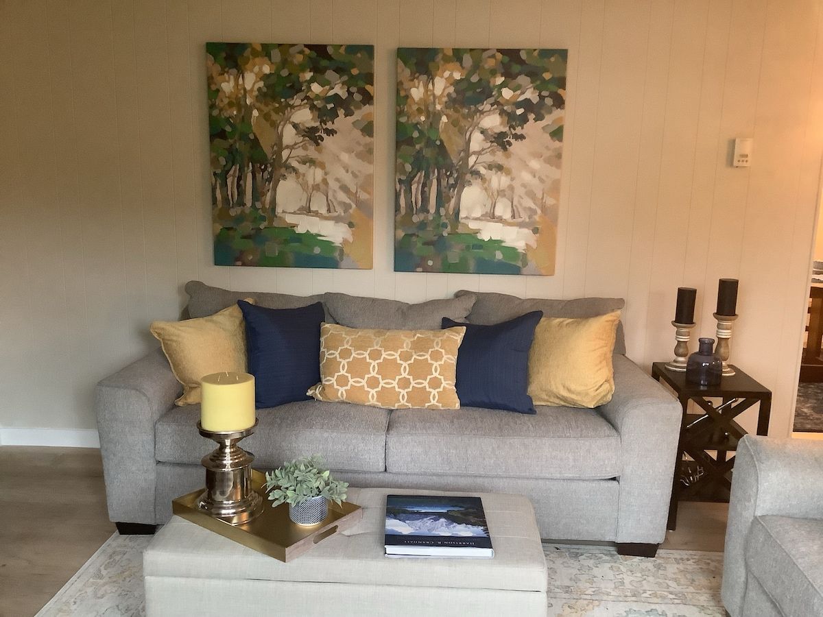 Uma sala de estar com um sofá cinza, almofadas estampadas, uma mesa pequena com uma vela e duas pinturas na parede.