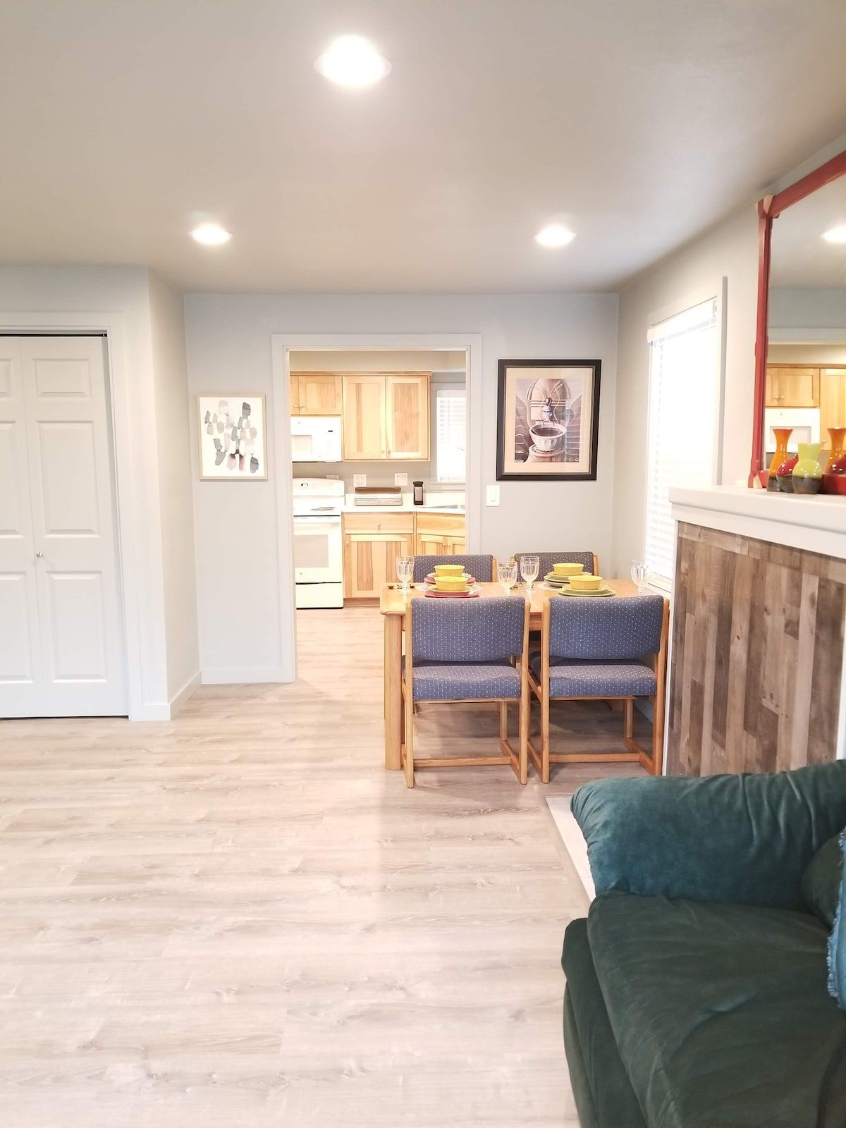 A dining area with a table set for two, a kitchen in the background, and a green sofa in the foreground.