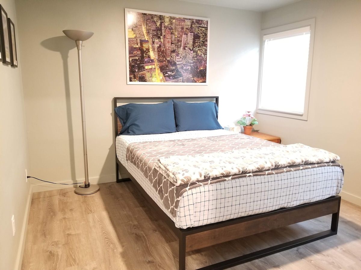 Quarto com cama, luminária de chão, pintura de paisagem urbana, janela e pequena mesa com planta.