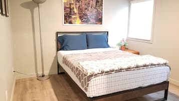 Une chambre à coucher avec un lit, une lampe sur pied, un tableau représentant un paysage urbain, une fenêtre et une petite table avec une plante.