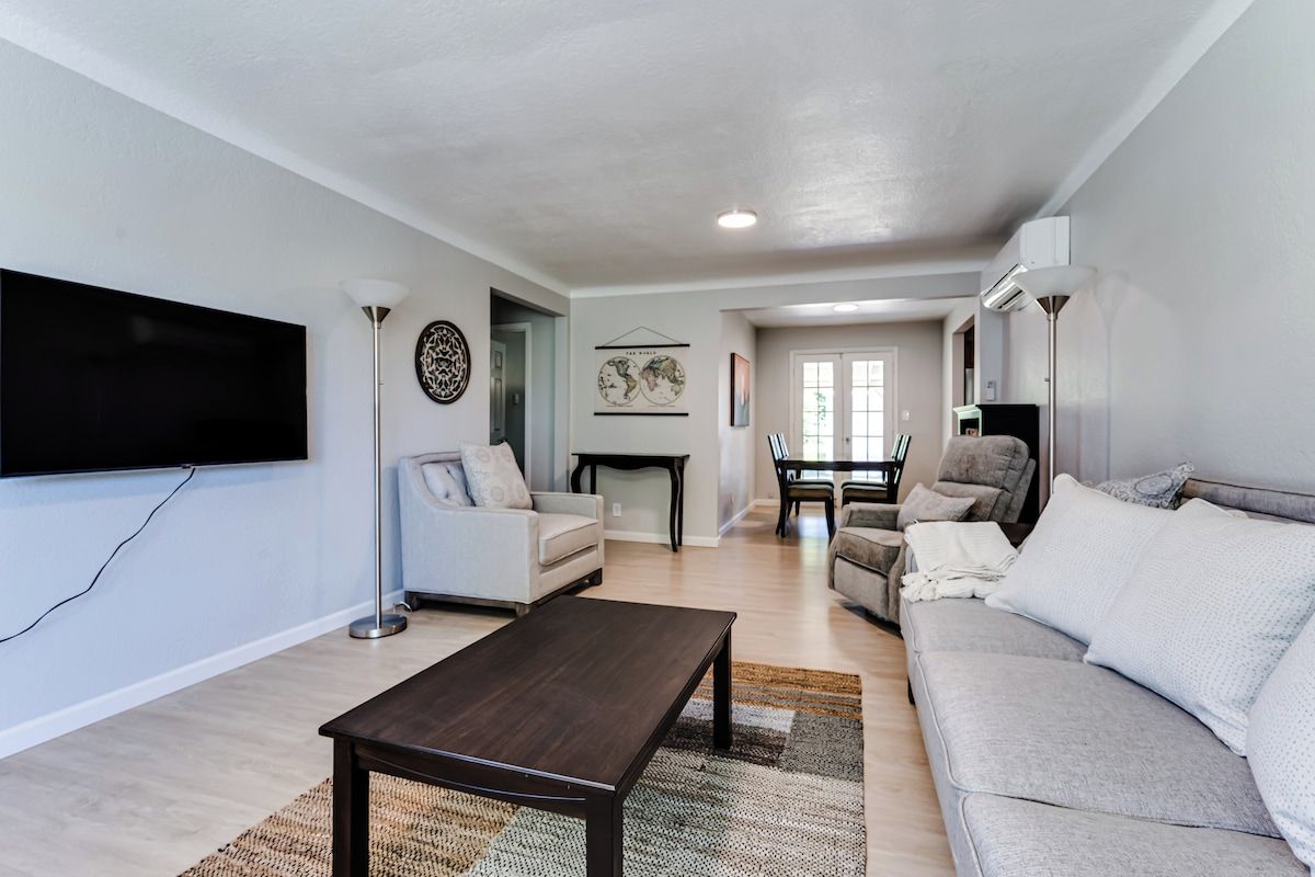 A living room with a flat-screen TV, a dark wooden coffee table, a grey sofa, and a white armchair.