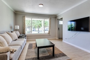 A living room with a sofa, a coffee table, a flat-screen TV mounted on the wall, and a window with curtains. - Central Eugene Home: Spacious Yard, Free Parking (Eugene)