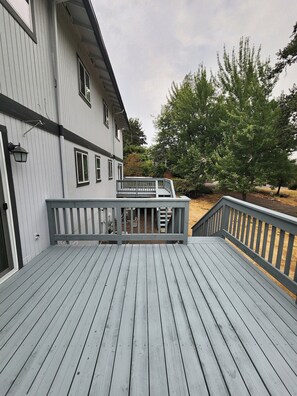 En träveranda med ett räcke, en byggnad med flera fönster och ett träd i bakgrunden.