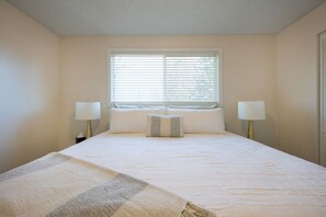 A hotel room with a large bed, two bedside lamps, a window with blinds, and a patterned throw blanket. - West Hills Retreat with Reclining Couch & King Bed (Eugene)