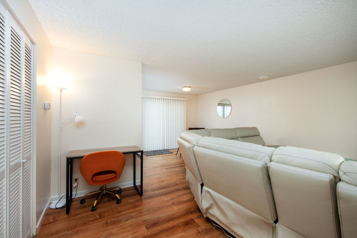A room with a white sofa, a desk with an orange chair, and a round mirror on the wall.