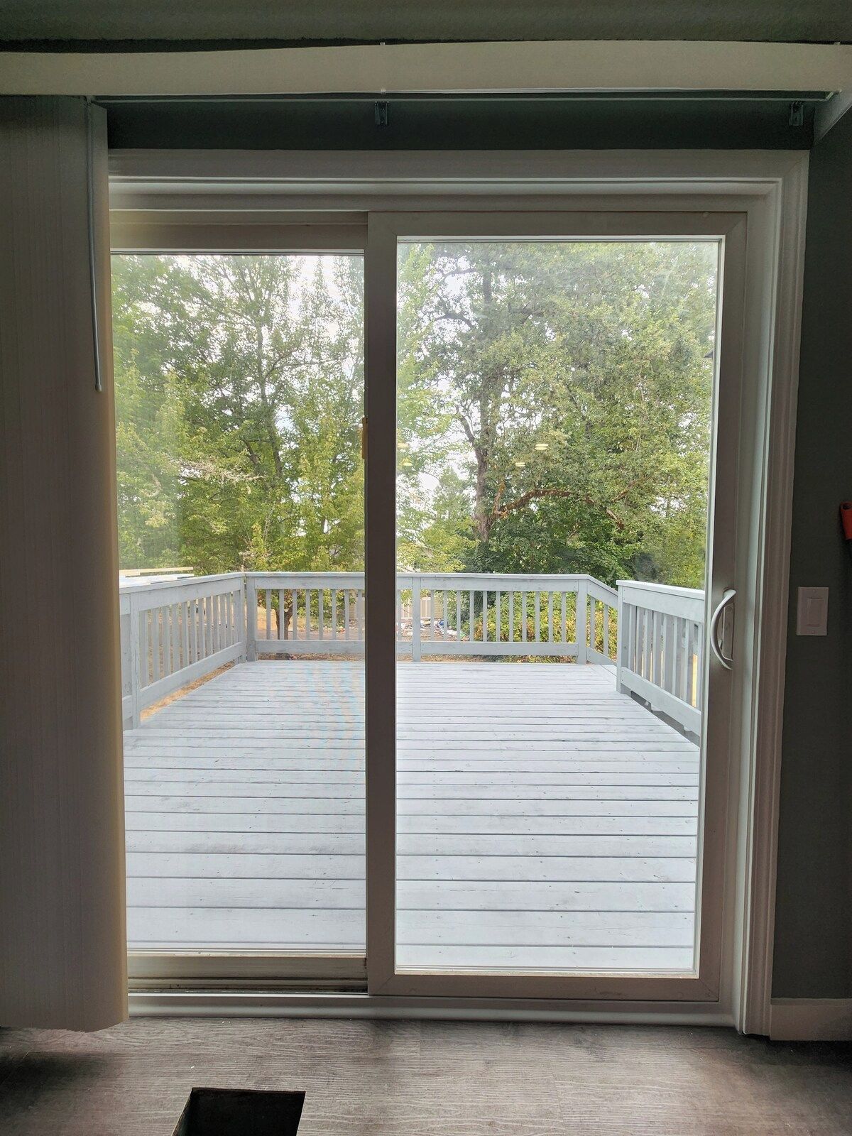 Vista a través de un vidrio corredero hacia una terraza de madera con barandilla blanca, que da a una zona verde y llena de árboles.