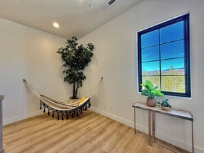 Ein Zimmer mit einer Hängematte, einem Blumentopf, einem Fenster mit Ausblick und einem kleinen Tisch mit weiteren Pflanzen.