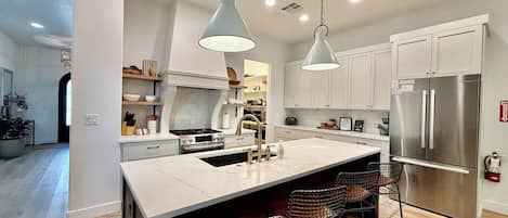 Una cocina moderna con una isla central, iluminación colgante, electrodomésticos de acero inoxidable y vistas al área de comedor.