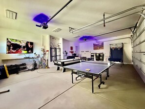 A room with two ping pong tables, a ceiling-mounted projector, and a wall-mounted television. - Hollow Sands Basecamp | Hot Tub | Pool | Fire Pit (Hurricane)