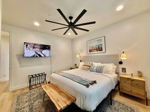 A bedroom with a bed, bedside tables, a bench, a ceiling fan, a TV, and wall-mounted lamps. - Hollow Sands Guest Suite | Hot Tub | Pool (Hurricane)