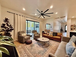 A modern living room with a ceiling fan, sofas, armchairs, a coffee table, and a large window with curtains. - Hollow Sands Retreat | Hot Tub | Pool | Duplex (Hurricane)