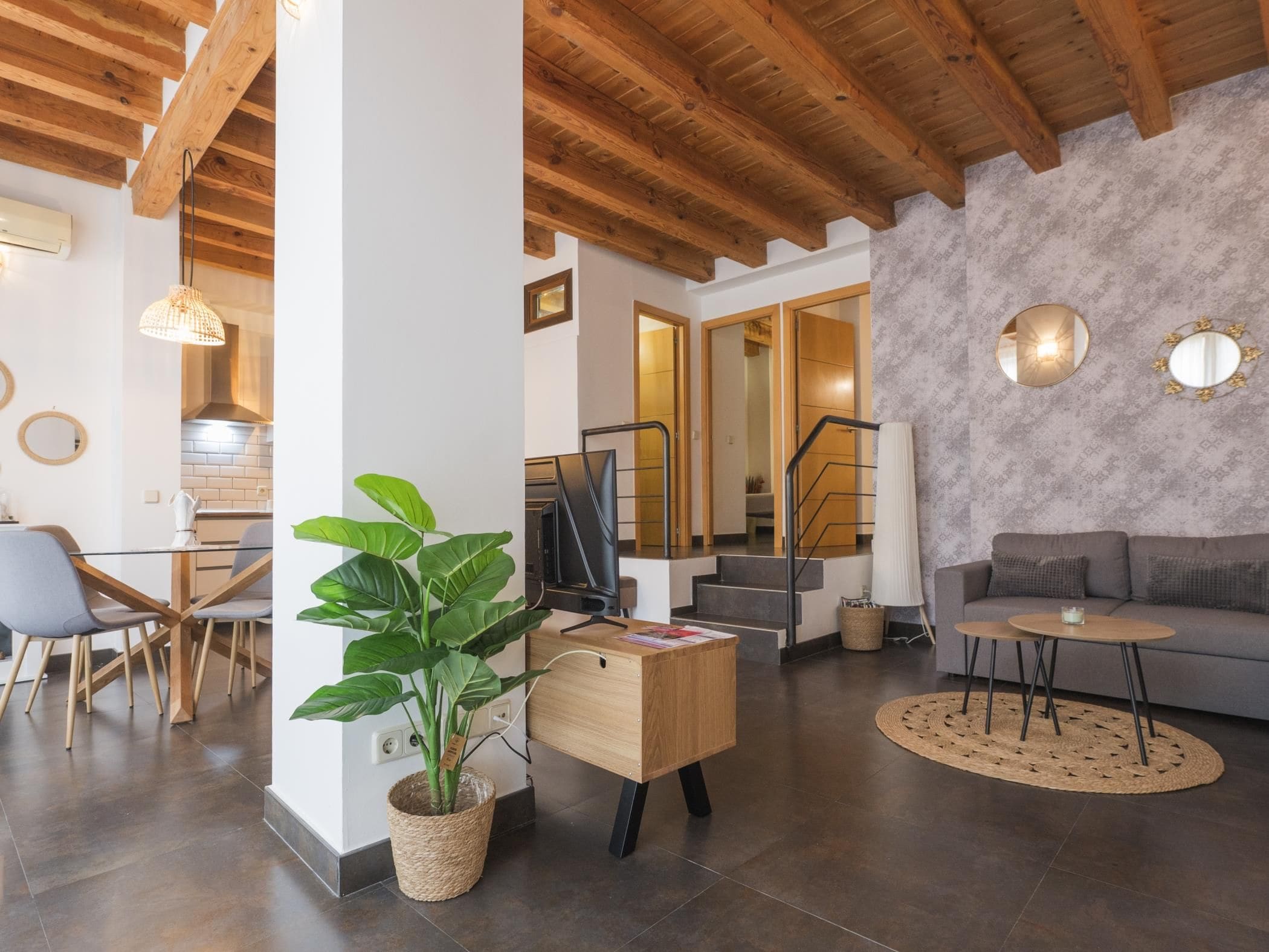 A modern interior with a wooden ceiling, a dining area with chairs, a sofa, a TV on a wooden stand, and a staircase.