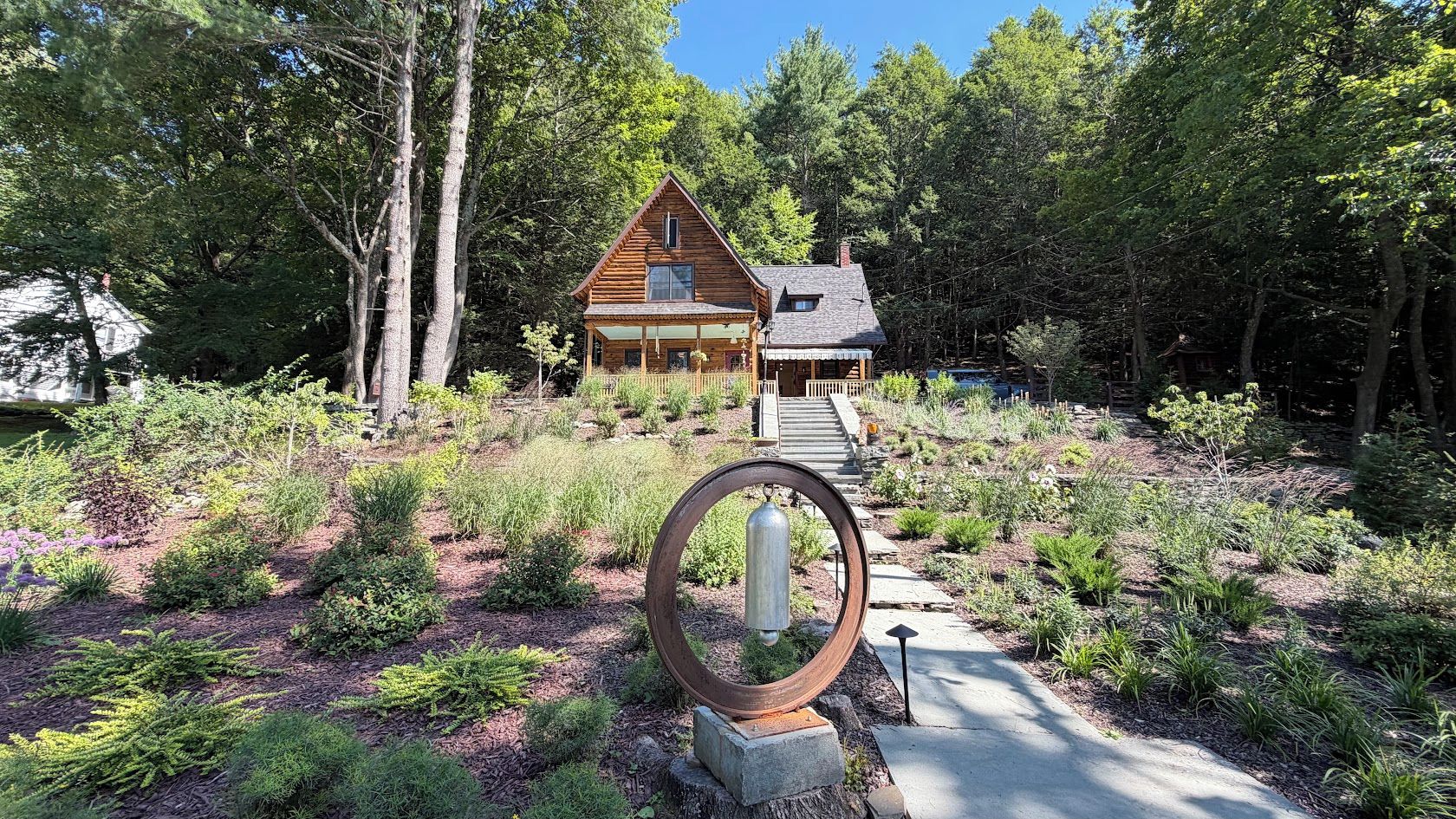A cabin with a porch, surrounded by a forested area and a garden with a metal sculpture.