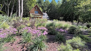 Una cabaña de madera rodeada por un jardín con flores moradas y diversos arbustos.