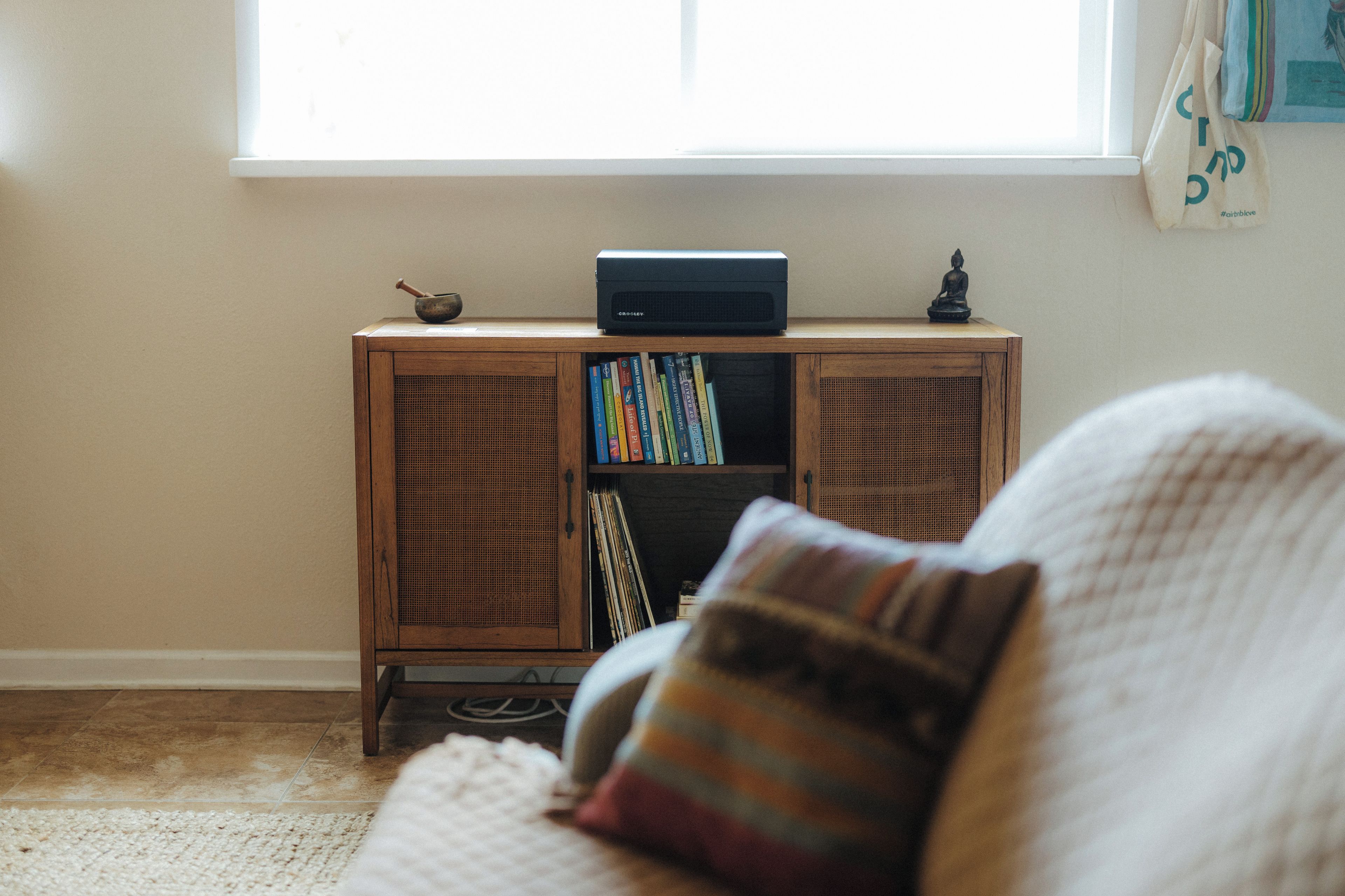 A room with a wooden cabinet containing books and a radio, a window with a view, and a couch with a striped pillow.