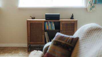 A room with a wooden cabinet containing books and a radio, a window with a view, and a couch with a striped pillow.