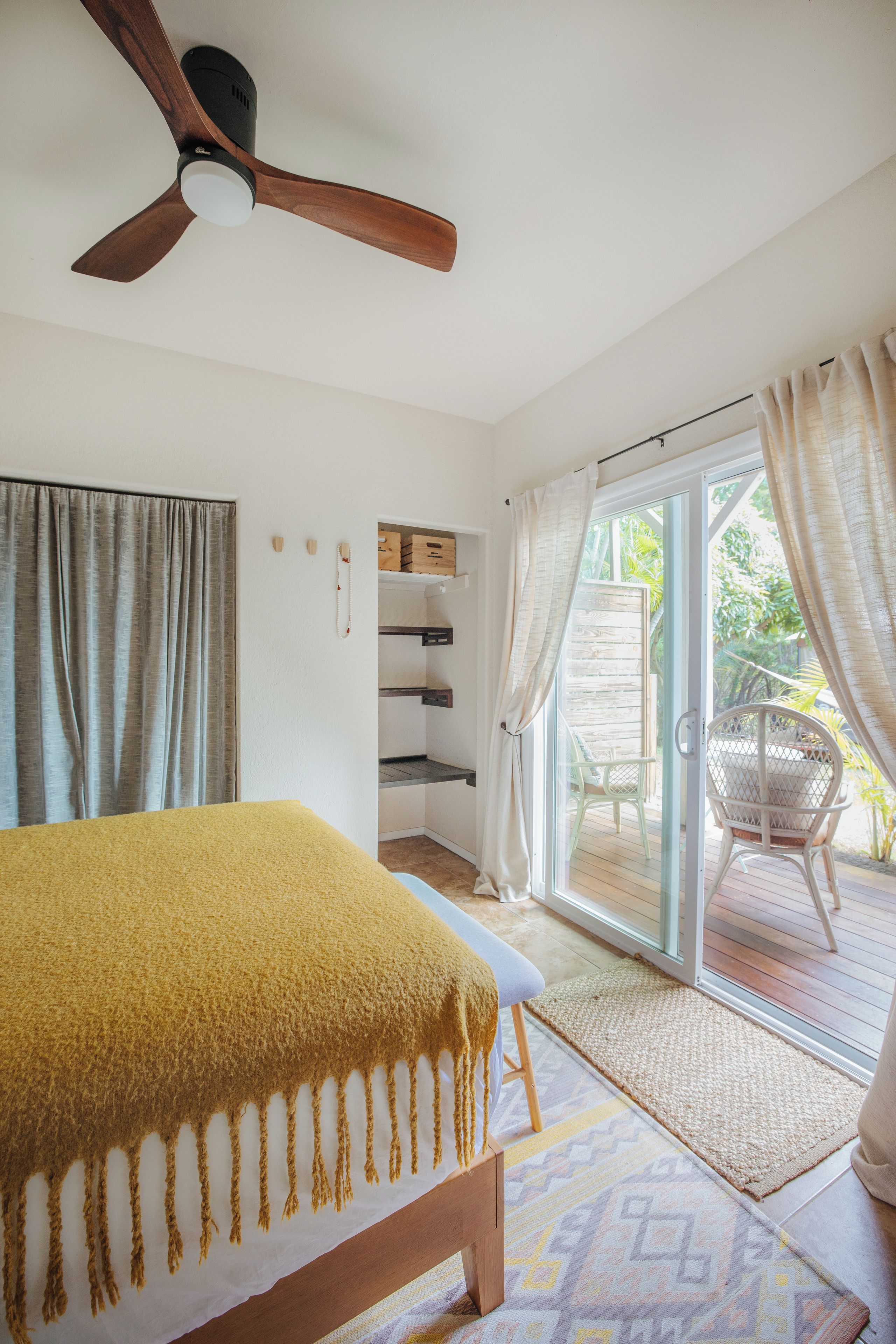 A bedroom with a bed, a ceiling fan, a sliding glass door leading to a patio with a chair, and a small shelf with boxes.