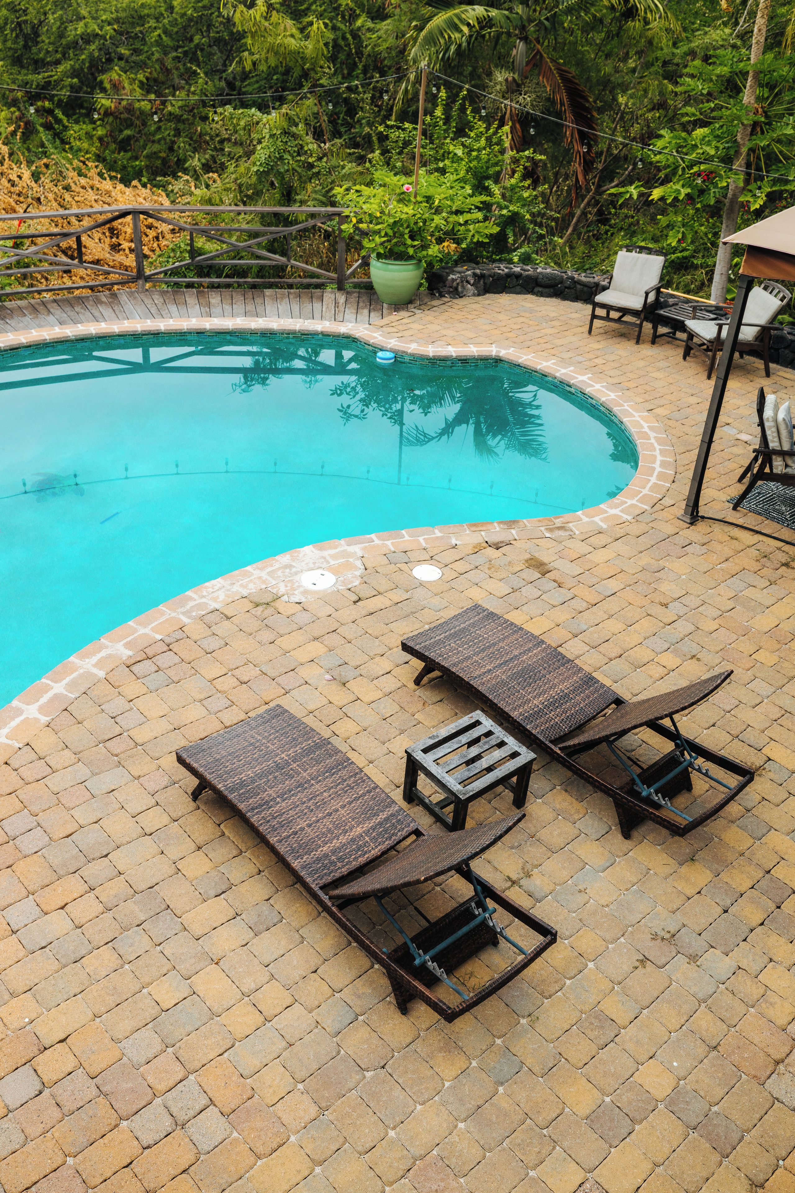 A swimming pool with a curved edge, surrounded by a brick patio. There are two lounge chairs and a small table on the patio.