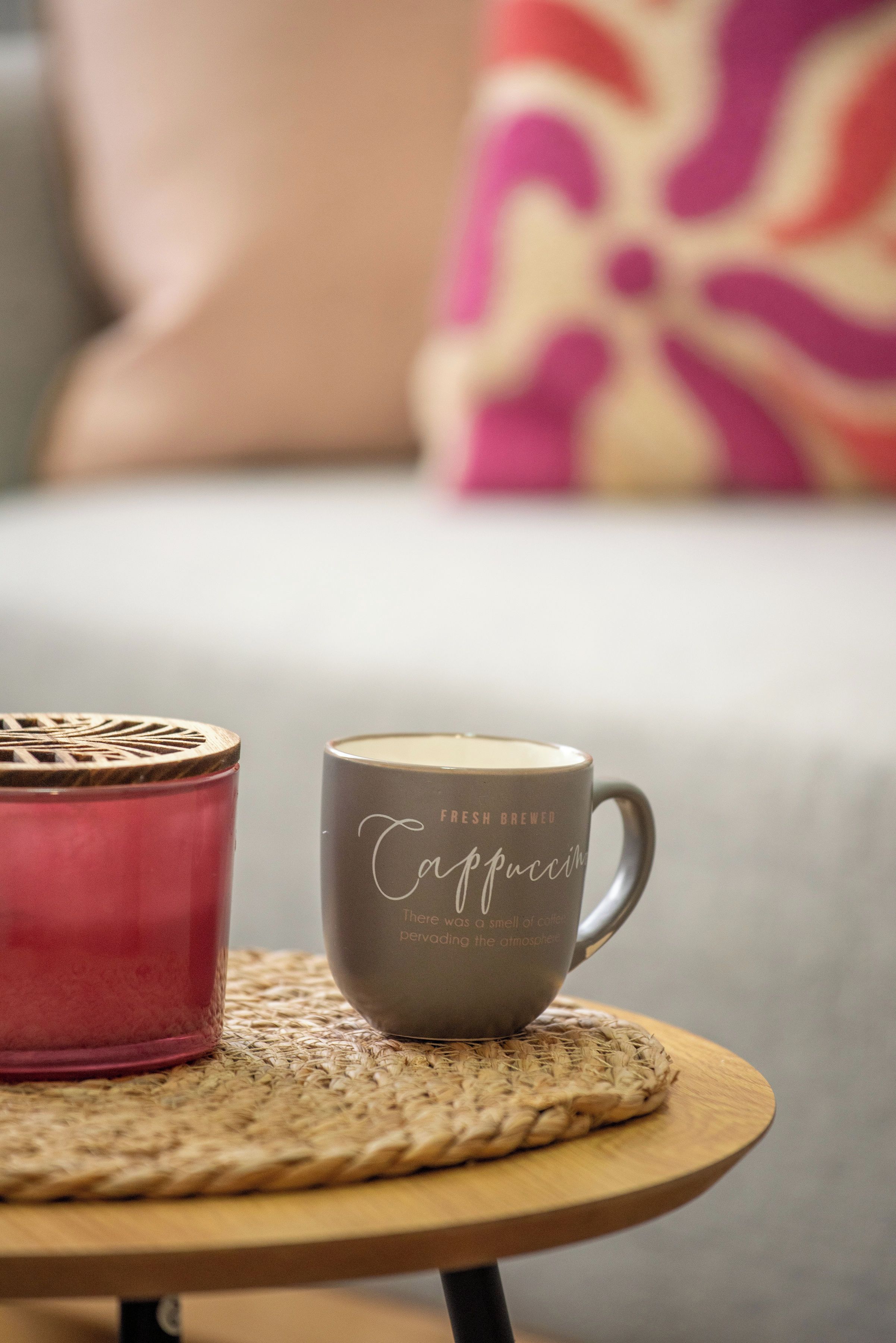 A cozy setting with a mug on a woven placemat, a candle, and a cushion in the background.