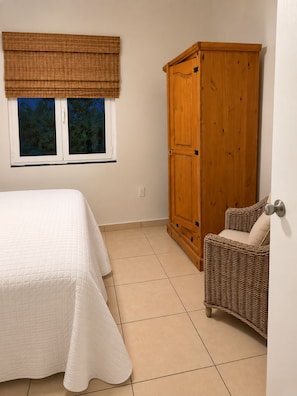 Ein Schlafzimmer mit einem Bett, einem Kleiderschrank, einem Korbsessel und einem Fenster mit Blick auf Bäume.