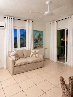 Ein Wohnzimmer mit einem beigefarbenen Sofa, Sternpilzen als Kissen, einem Strandbild an der Wand, einem Deckenventilator und Blick durch eine Glastür.