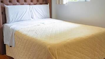 Uma cama de solteiro com cabeceira, uma janela com cortinas e piso de madeira.