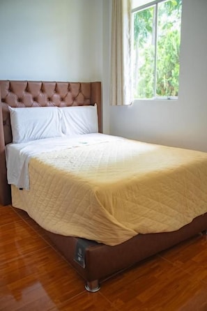 Una cama individual con cabecera, una ventana con cortinas y piso de madera.