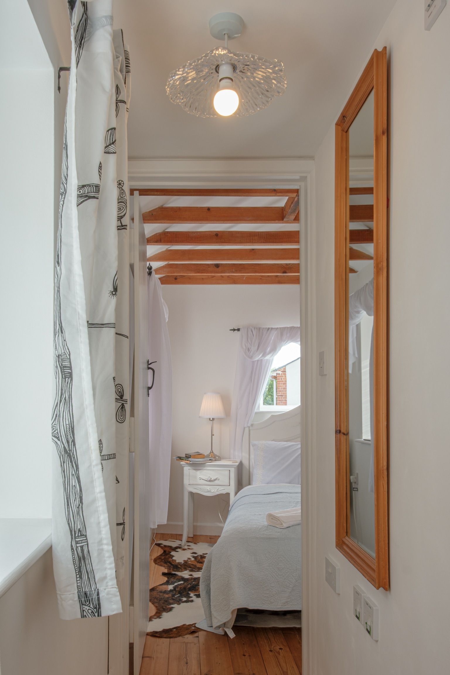 A narrow hallway leading to a bedroom with a bed, a nightstand, a lamp, and a mirror.
