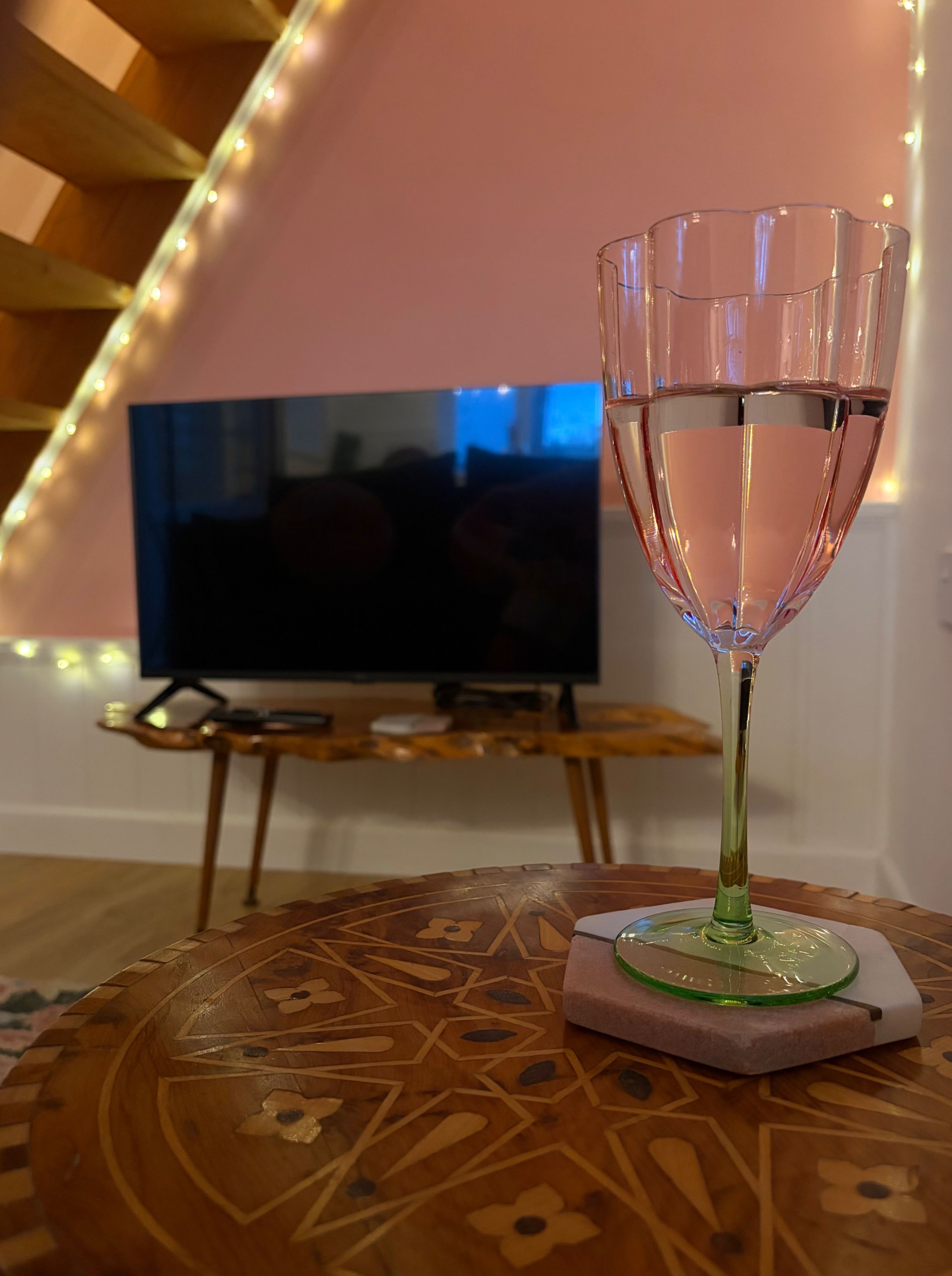 A glass of water on a coaster with a geometric pattern, a television in the background, and a staircase with string lights.