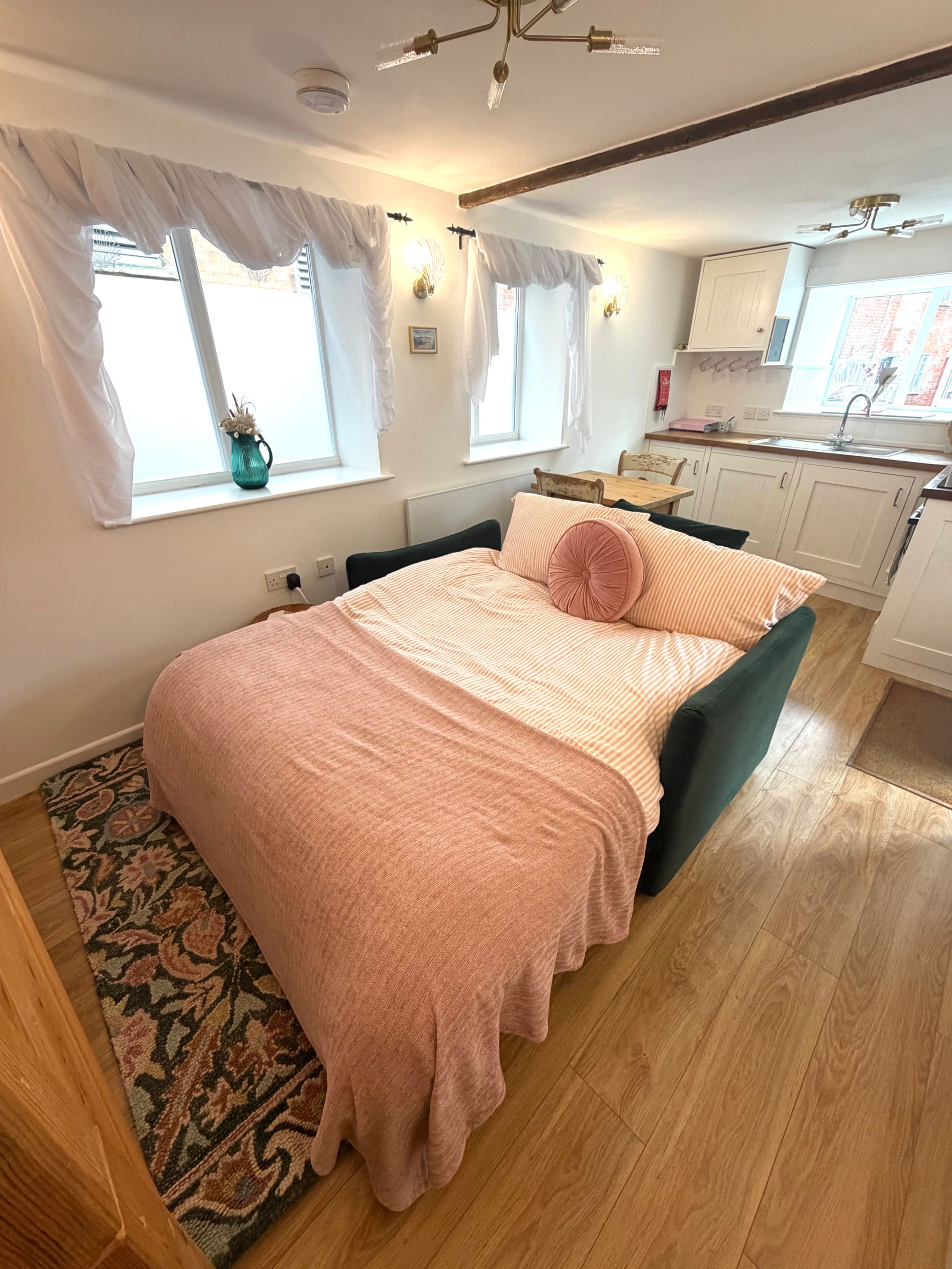 Quarto com cama, janela, uma mesinha e uma kitchenette ao fundo.