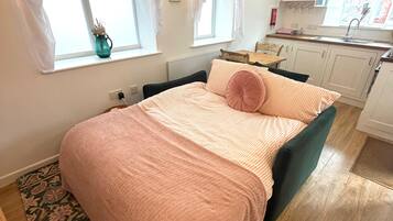 Quarto com cama, janela, uma mesinha e uma kitchenette ao fundo.