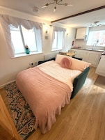 Un dormitorio con cama, ventana, una mesita y una kitchenette al fondo.