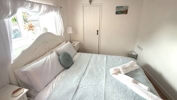 Un dormitorio con cama, mesita de noche, lámpara, ventana con cortinas, puerta y un pequeño cuadro en la pared.