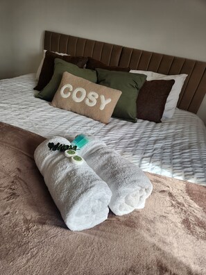 Una cama bien tendida con cabecera, almohada con la palabra 'COSY' y dos toallas enrolladas con un adorno de hoja verde.
