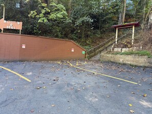 A parking area with yellow lines and scattered leaves, a brick wall, and a wooden staircase leading up. - Cozy Cabin Nestled on Infamous Ski Mountain Rd. (Gatlinburg)