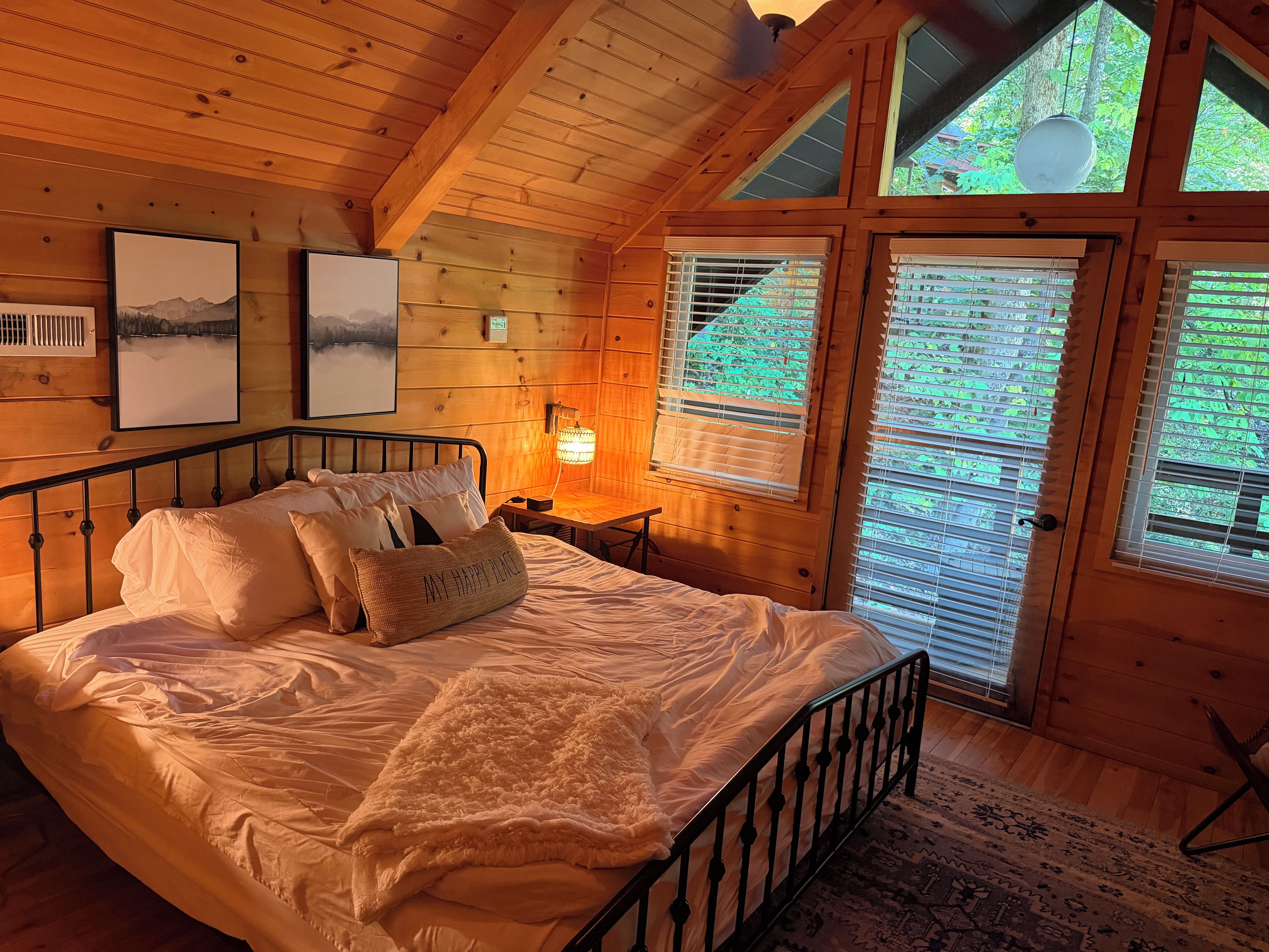 A wooden room with a bed, a nightstand, a lamp, and a window with blinds.
