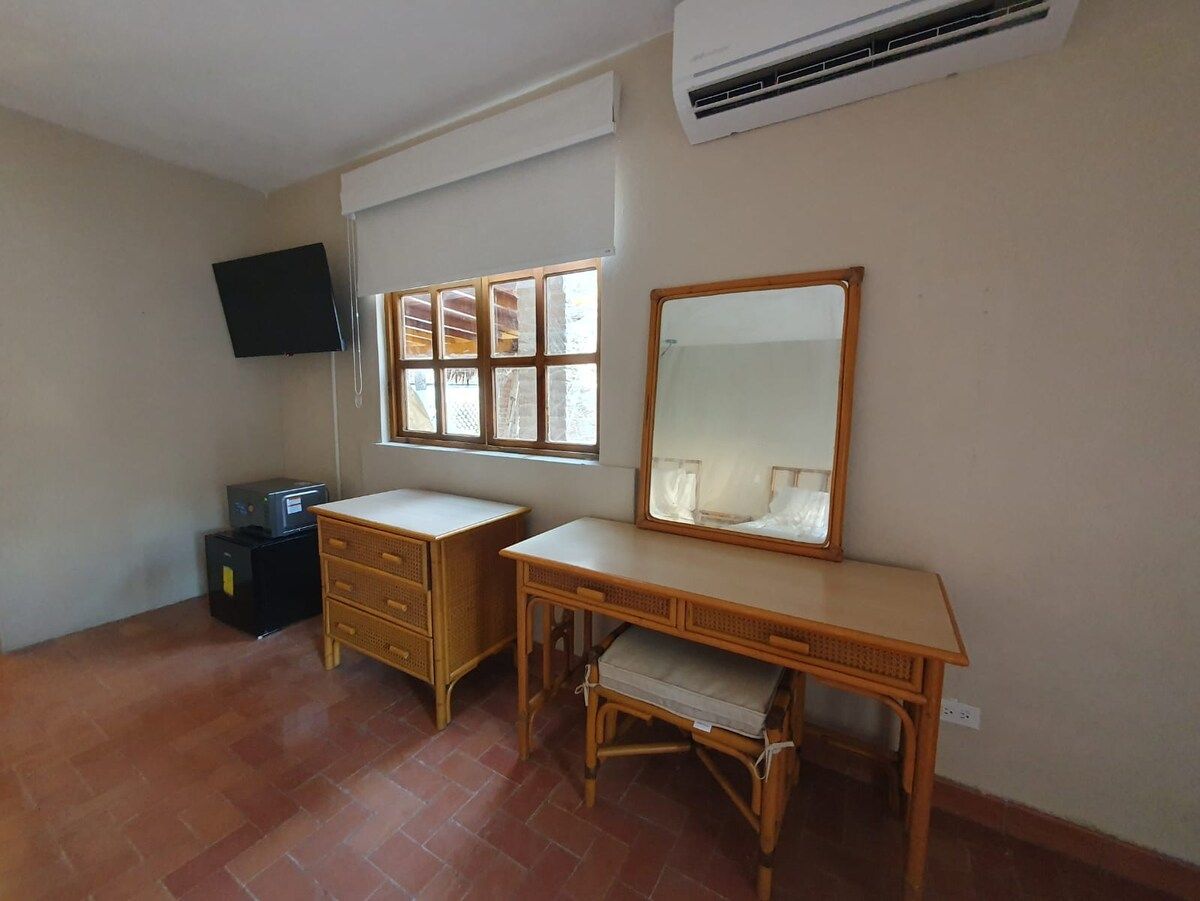 Una habitación con escritorio, silla, televisor montado en la pared y ventana con persianas.