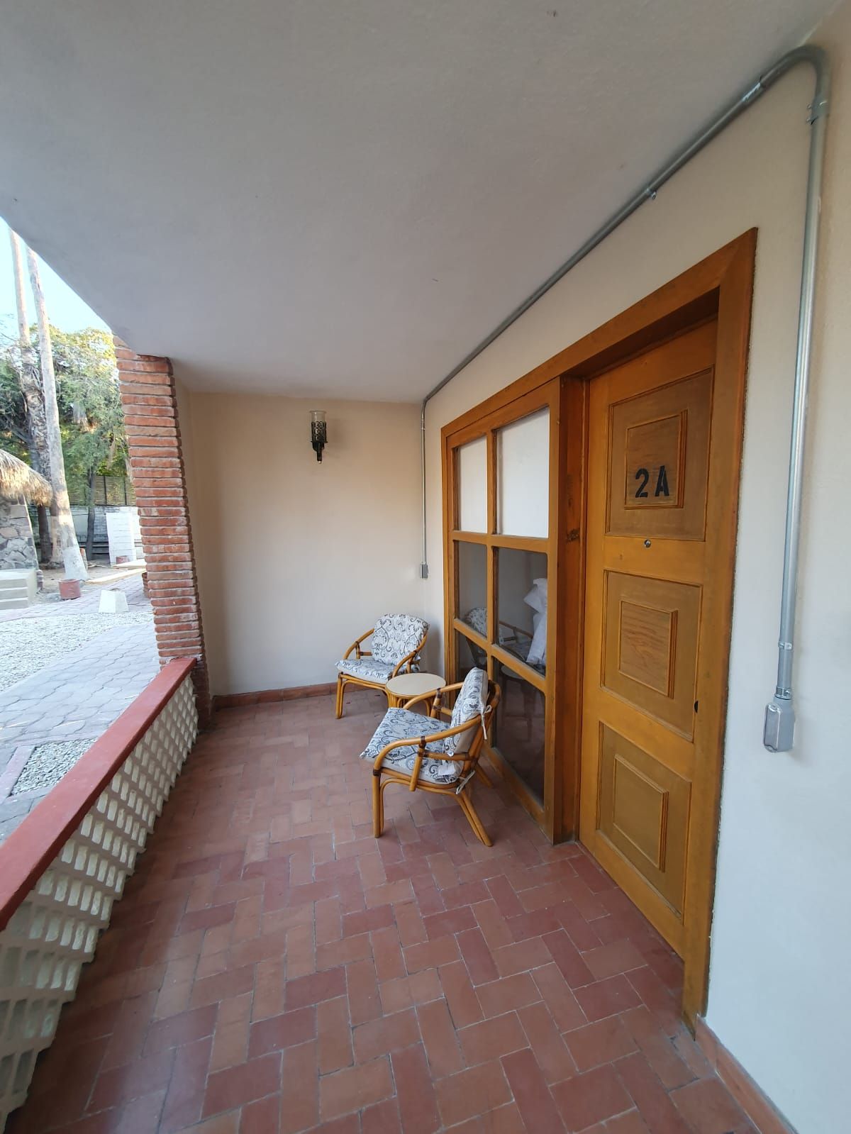 Una habitación con suelo de baldosas, dos sillones de mimbre, una puerta de madera con un número y una lámpara fijada a la pared.
