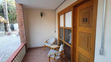 A room with a tiled floor, two wicker chairs, a wooden door with a number, and a wall-mounted light fixture.