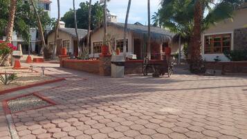 A courtyard with hexagonal paving, palm trees, and buildings with thatched roofs.