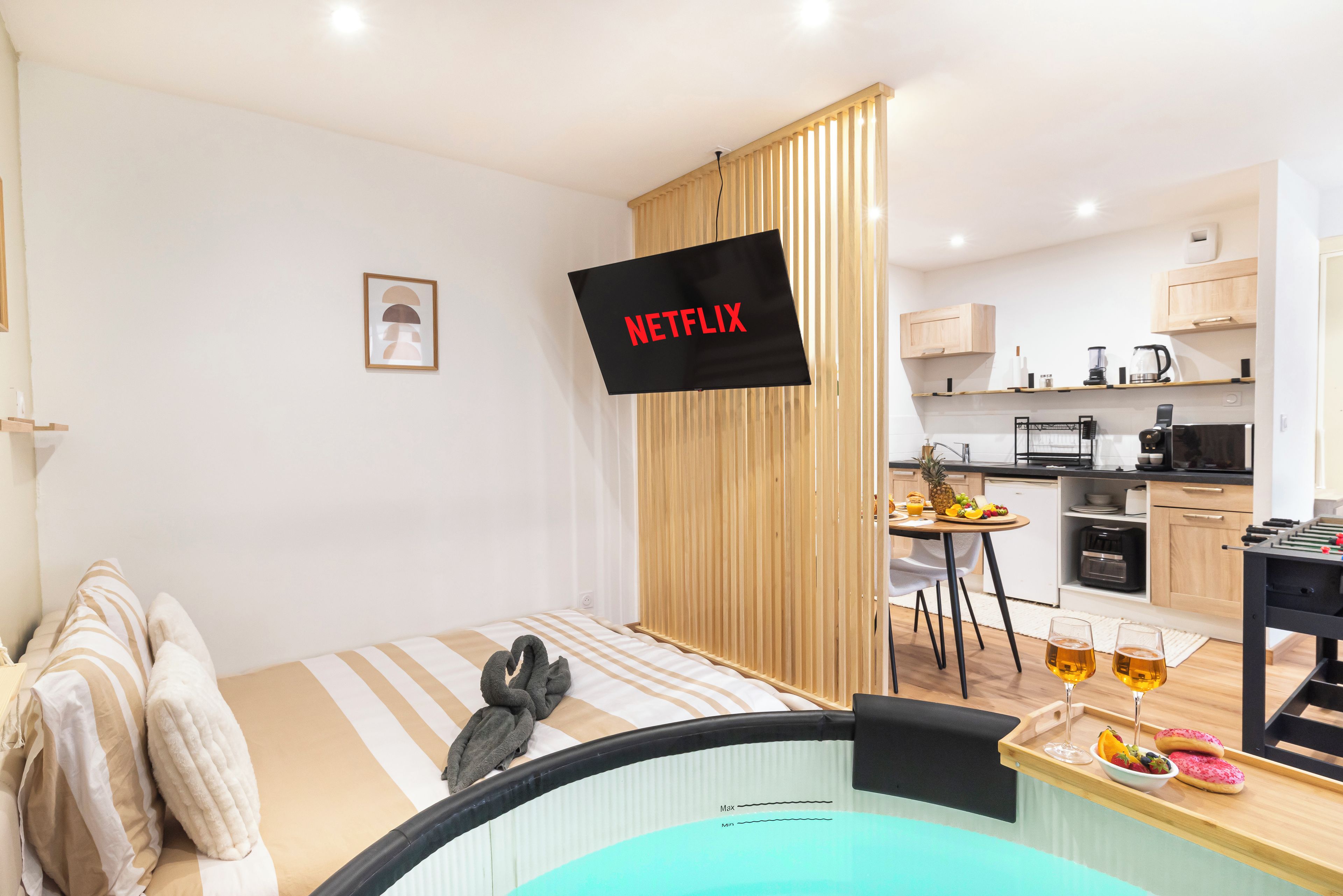 A modern hotel room with a bed, a small kitchenette, a dining area, and a TV screen displaying 'NETFLIX'.