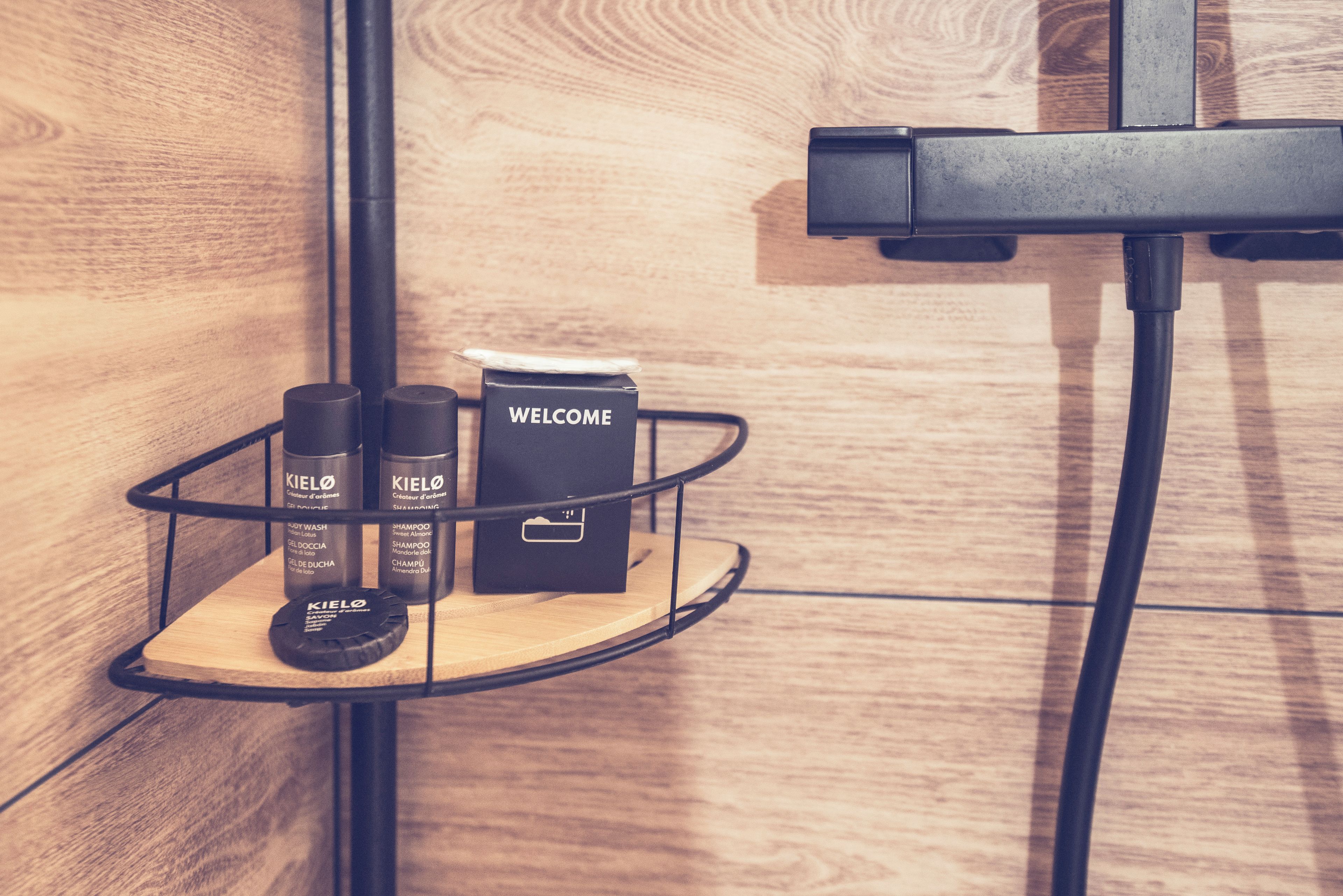 A shower area with a black metal shelf holding toiletries and a welcome box.