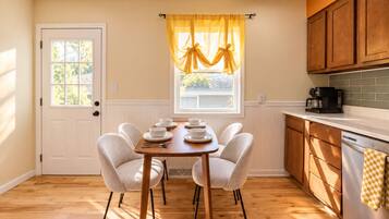 A dining area with a wooden table and chairs, a window with yellow curtains, and a kitchen with wooden cabinets and a coffee maker.