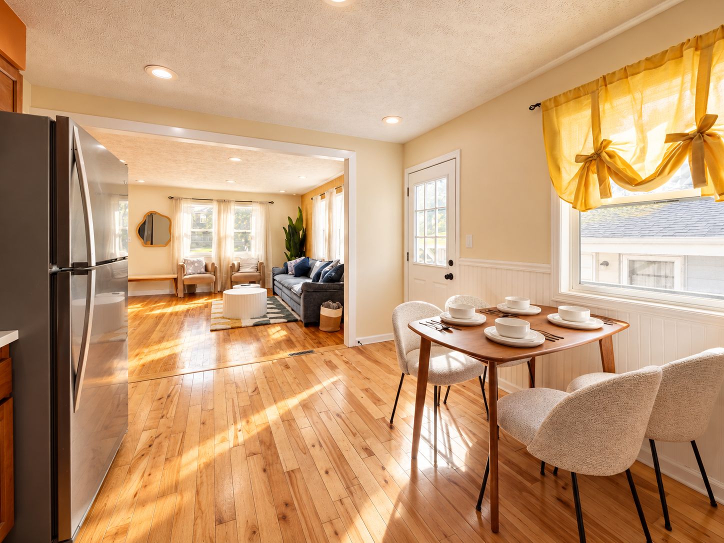 Una cocina moderna con piso de madera, un comedor con una mesa preparada para cuatro personas, y una sala de estar con un sofá y sillones.